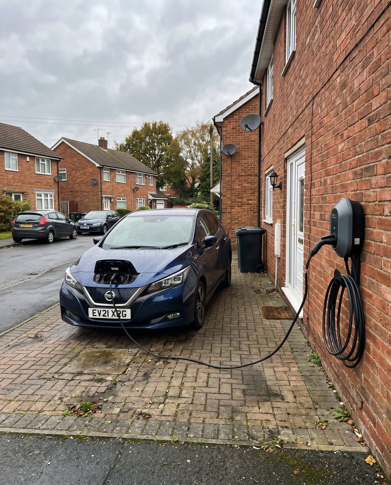 Blue electric car charging at a home charging station in a residential driveway with brick houses