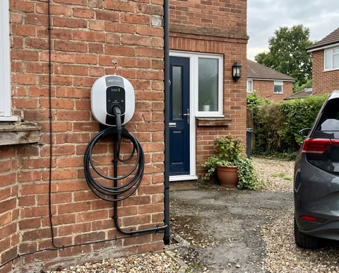 White and black electric car charging station mounted on brick wall of residential house with driveway and parked vehicle visible