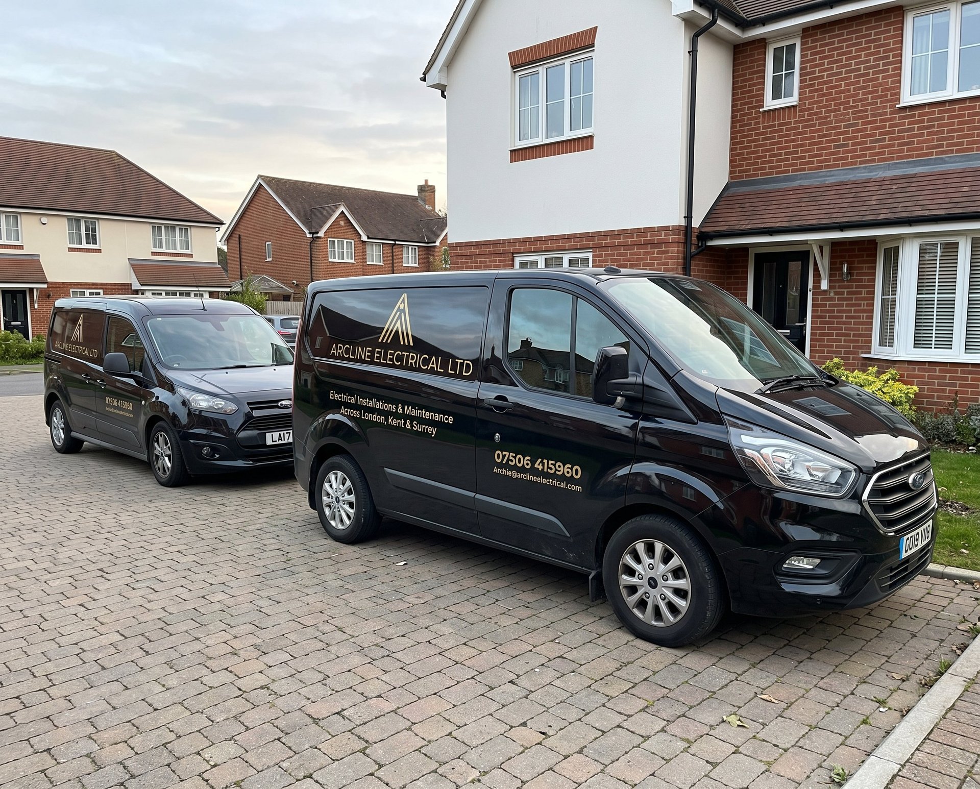 Two black commercial vans parked on a brick driveway in front of a two-story brick house