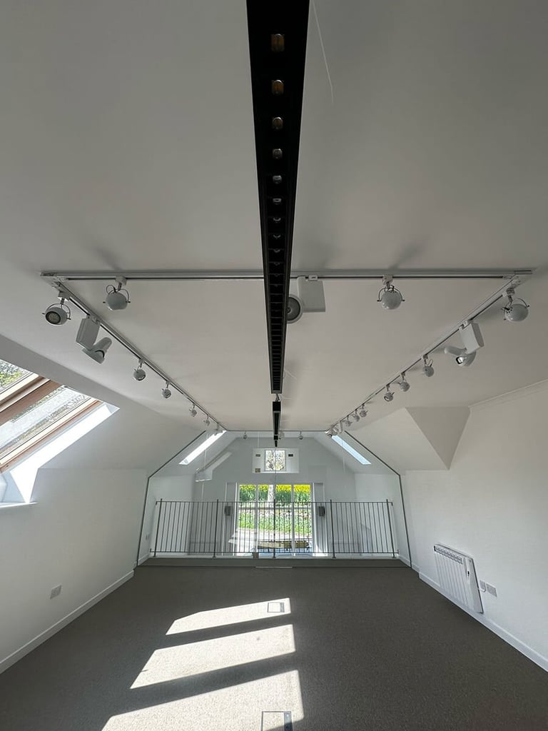 Empty gallery or studio space with high ceilings, track lighting, skylights, and a central black beam, photographed from the entrance
