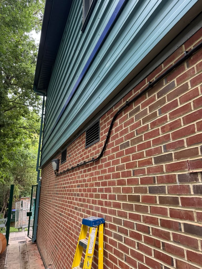 Red brick wall with metal gutter and downspout, yellow ladder leaning against wall, outdoor residential area with trees