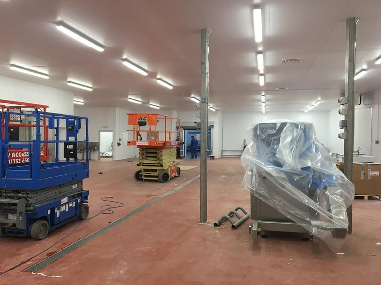 Warehouse or factory floor with industrial equipment including blue scissor lift and orange machinery, with plastic-wrapped items and metal support columns