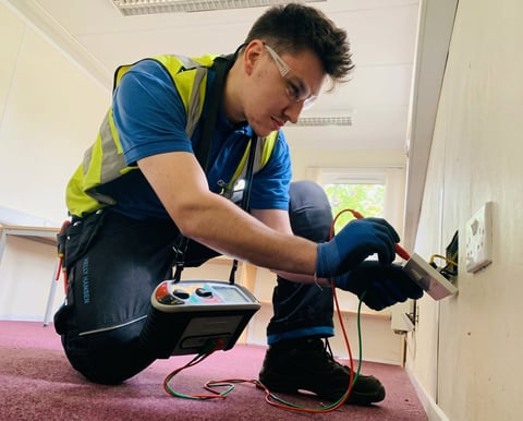 Electrician in yellow safety vest and glasses testing electrical outlet with multimeter on pink carpet indoors